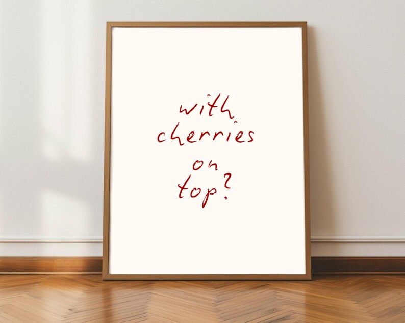 Puede incluir: Un p&oacute;ster enmarcado con la frase "with cherries on top?" en rojo manuscrito sobre un fondo blanquecino. El marco es de color marr&oacute;n claro. El p&oacute;ster se muestra contra una pared blanca y un suelo de madera.