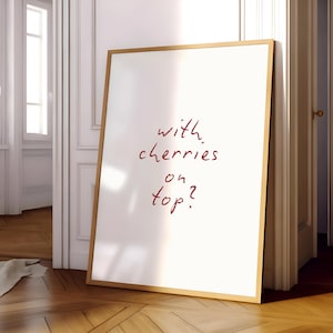 Puede incluir: Un cuadro blanco enmarcado con el texto manuscrito "with cherries on top?" en rojo. El marco es de color marr&oacute;n claro. El cuadro est&aacute; apoyado contra una pared blanca y un suelo de madera.