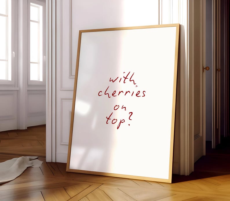 Puede incluir: Un cuadro blanco enmarcado con el texto manuscrito "with cherries on top?" en rojo. El marco es de color marr&oacute;n claro. El cuadro est&aacute; apoyado contra una pared blanca y un suelo de madera.