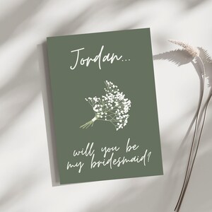 May include: A green card with white text that reads "Jordan... will you be my bridesmaid?" and a small white flower bouquet.