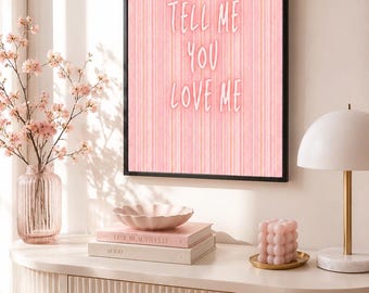 May include: A rectangular wall art print in a black frame featuring a pink striped background with the white text "TELL ME YOU LOVE ME" centered. The print is displayed above a white desk with a pink stool, a pink vase with flowers, and books.
