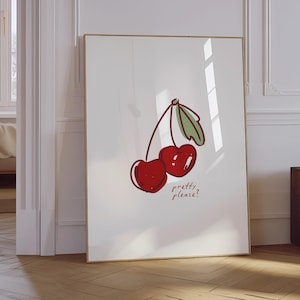 Puede incluir: Una impresi&oacute;n art&iacute;stica enmarcada con una ilustraci&oacute;n caprichosa de dos cerezas rojas con hojas y tallos verdes. El texto "pretty please?" est&aacute; escrito debajo. La obra de arte se exhibe en una habitaci&oacute;n luminosa con suelo de madera.