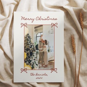 Puede incluir: Una tarjeta de Navidad con una foto de una familia de pie frente a un &aacute;rbol de Navidad decorado. La tarjeta tiene un borde rojo con lazos en cada esquina. El texto "Feliz Navidad, los Daniels, 2024" est&aacute; impreso en la tarjeta.