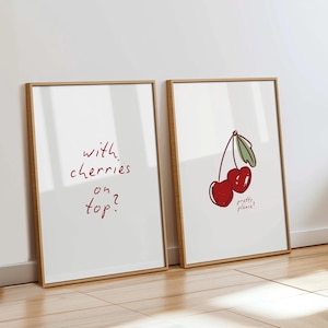 Puede incluir: Dos impresiones art&iacute;sticas enmarcadas con fondos blancos y marcos de madera. Una impresi&oacute;n tiene el texto "with cherries on top?" en rojo. La otra presenta una ilustraci&oacute;n de cerezas con el texto "pretty please?"