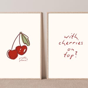 Puede incluir: Dos impresiones art&iacute;sticas enmarcadas con fondo crema. Una impresi&oacute;n presenta una ilustraci&oacute;n de cerezas con el texto "pretty please?". La otra dice "with cherries on top?" en escritura roja.