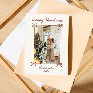 Puede incluir: Una tarjeta de Navidad blanca con un borde rojo y lazos rojos. La tarjeta presenta una foto de una familia de pie frente a un &aacute;rbol de Navidad. El texto en la tarjeta dice "Feliz Navidad, los Daniels, 2024".