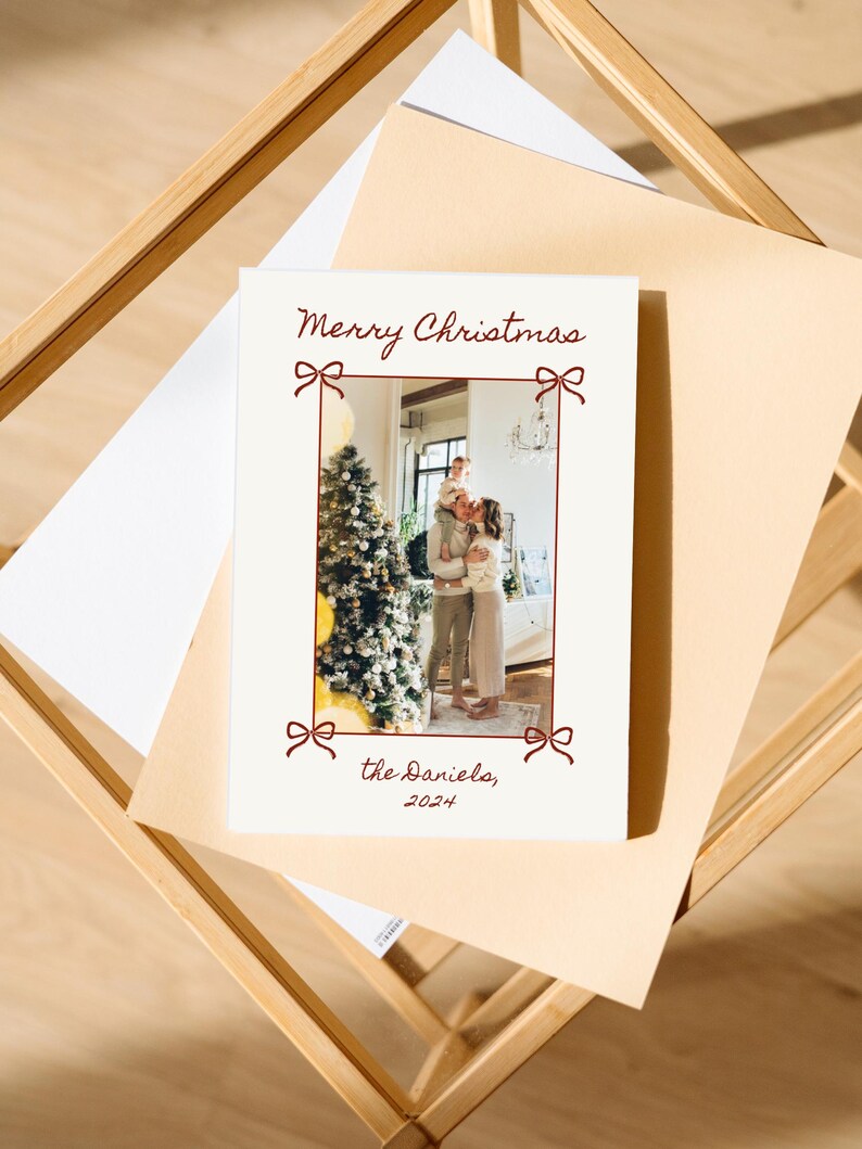 Puede incluir: Una tarjeta de Navidad blanca con un borde rojo y lazos rojos. La tarjeta presenta una foto de una familia de pie frente a un &aacute;rbol de Navidad. El texto en la tarjeta dice "Feliz Navidad, los Daniels, 2024".