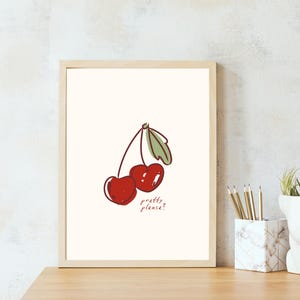 Puede incluir: L&aacute;mina art&iacute;stica enmarcada con una ilustraci&oacute;n de dos cerezas rojas con hojas verdes y el texto "pretty please?". La obra de arte est&aacute; sobre un fondo crema dentro de un marco de madera clara.