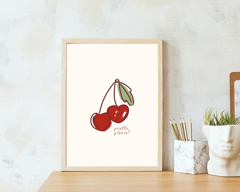 Puede incluir: L&aacute;mina art&iacute;stica enmarcada con una ilustraci&oacute;n de dos cerezas rojas con hojas verdes y el texto "pretty please?". La obra de arte est&aacute; sobre un fondo crema dentro de un marco de madera clara.