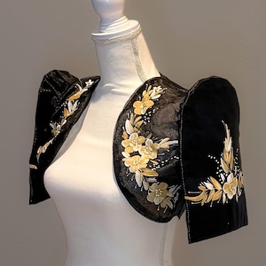 May include: Black bolero jacket with floral embroidery in gold and white. The jacket features puffed sleeves and is displayed on a white mannequin. The floral design is concentrated on the shoulders and sleeves.