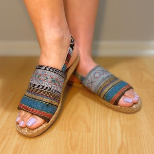 May include: A pair of open-toe sandals with a woven fabric design in shades of blue, brown, and red. The sandals have a jute rope sole and a strap around the ankle. The sandals are on a wooden floor.