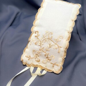 May include: Ivory-colored rectangular sachet with a scalloped edge and gold floral embroidery. Two ivory ribbons are attached at the bottom. The sachet is made of a delicate fabric, and the design is elegant and ornate.