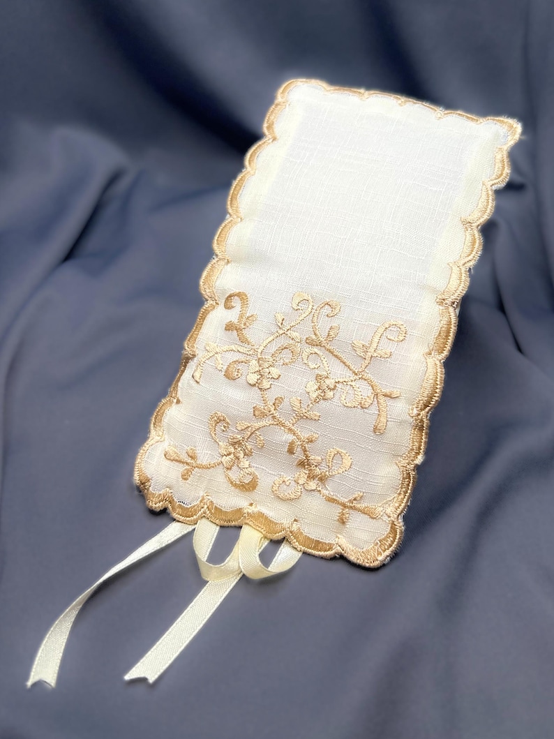 May include: Ivory-colored rectangular sachet with a scalloped edge and gold floral embroidery. Two ivory ribbons are attached at the bottom. The sachet is made of a delicate fabric, and the design is elegant and ornate.