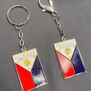 May include: Two keyrings displaying the flag of the Philippines. Each rectangular pendant has a red, white, and blue design with a gold sun and stars. The word "PILIPINAS" is printed at the top. One keyring has a ring, the other a clasp.