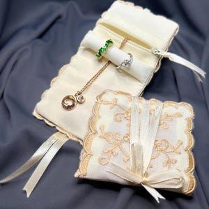Può includere: Astuccio portagioie color avorio con cerniera dorata, contenente un anello con pietra verde, un anello di diamanti e una collana d'oro con un ciondolo. È presente anche una pochette quadrata più piccola abbinata con un design in pizzo e chiusura a nastro.