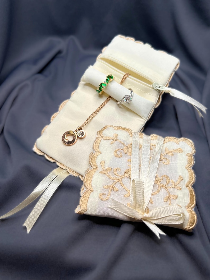 May include: Ivory jewelry roll with a gold zipper, holding a green gemstone ring, a diamond ring, and a gold necklace with a pendant. A smaller, matching square pouch with a lace design and ribbon closure is also present.