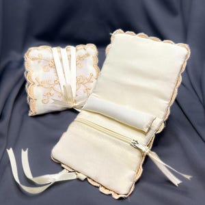 May include: Two ivory-colored jewelry cases. One is a square, tied with a ribbon, and the other is rectangular with a zipper and a small pillow inside. Both cases have a scalloped edge with a gold-colored trim.