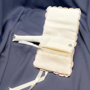 May include: Ivory-colored clutch with a scalloped edge and a small zippered pocket. The clutch has a matching ribbon and a small silver-colored clasp. The background is a dark blue fabric.