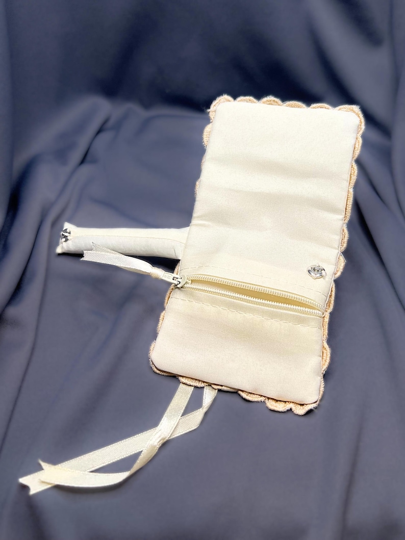 May include: Ivory-colored clutch with a scalloped edge and a small zippered pocket. The clutch has a matching ribbon and a small silver-colored clasp. The background is a dark blue fabric.