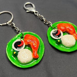 May include: Two keychains featuring miniature food replicas. Each keychain has a green plate with a piece of orange chicken, white rice, a small bowl of sauce, and a green garnish. The keychains have silver-colored key rings.
