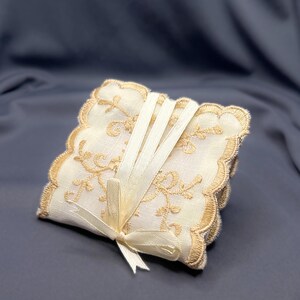 May include: A stack of square, off-white coasters with gold floral embroidery and scalloped edges. The coasters are tied together with a cream-colored ribbon and bow, creating an elegant and decorative set.
