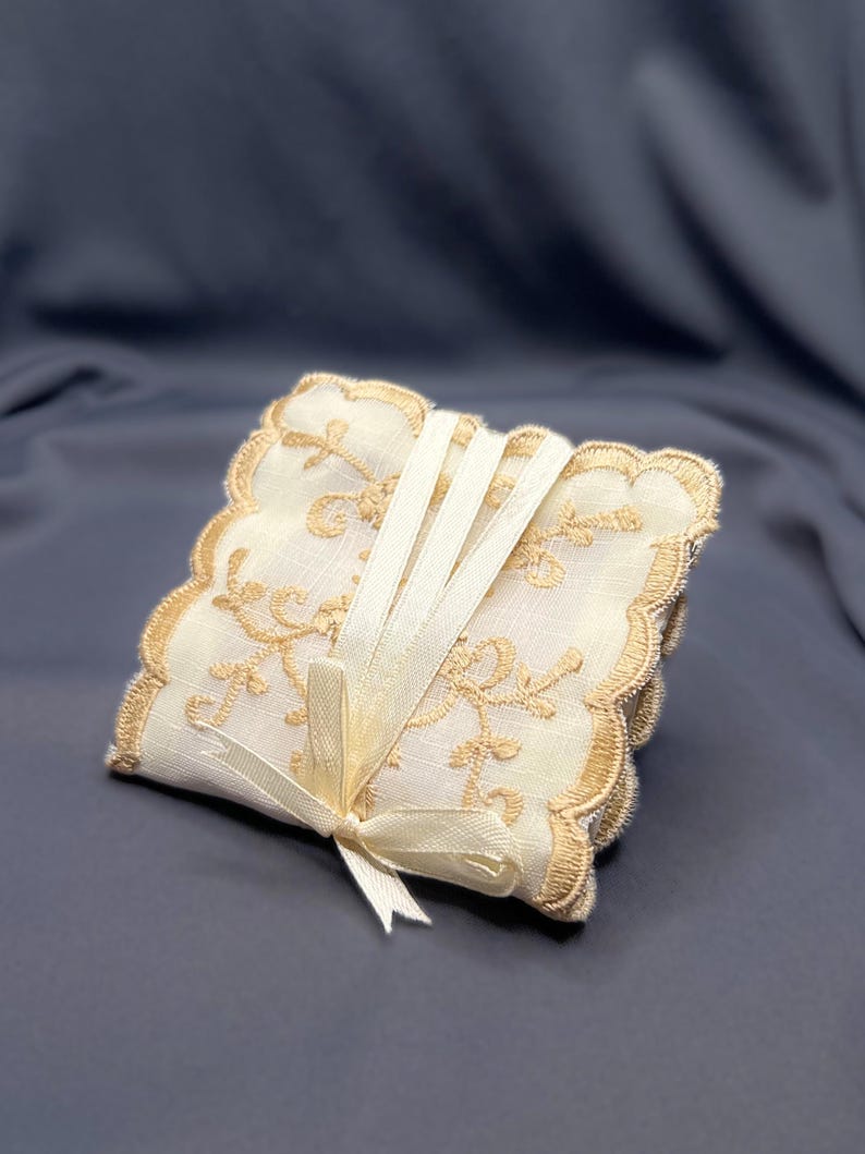 May include: A stack of square, off-white coasters with gold floral embroidery and scalloped edges. The coasters are tied together with a cream-colored ribbon and bow, creating an elegant and decorative set.