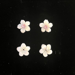 Puede incluir: Cuatro pendientes en forma de flor dispuestos en formación cuadrada. Los dos superiores tienen pétalos blancos con una piedra preciosa rosa en el centro, mientras que los dos inferiores tienen pétalos blancos con un pequeño punto metálico central.