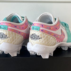 May include: Pair of white and pastel-colored Air Jordan cleats. The shoes feature a pink and teal design with sprinkles and the names "Brady" and "Irish". The Nike swoosh is pink. The soles are white with cleats.
