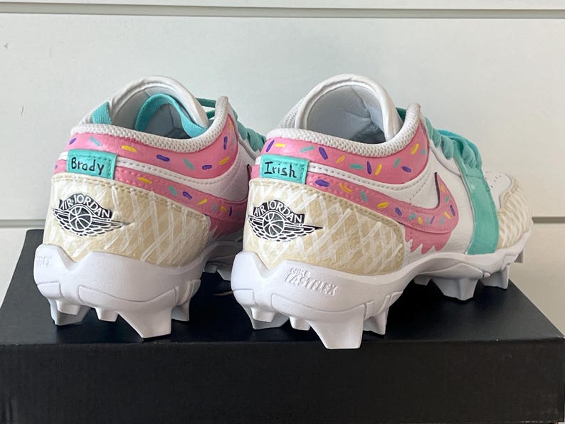 May include: Pair of white and pastel-colored Air Jordan cleats. The shoes feature a pink and teal design with sprinkles and the names "Brady" and "Irish". The Nike swoosh is pink. The soles are white with cleats.
