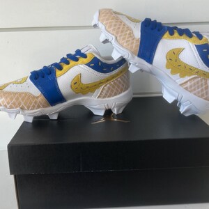 Puede incluir: Un par de tacos blancos, azules y amarillos con un dise&ntilde;o &uacute;nico. Los tacos presentan un patr&oacute;n similar a un gofre en la puntera y el tal&oacute;n, con un logotipo de Nike amarillo. Los zapatos est&aacute;n colocados encima de una caja negra.