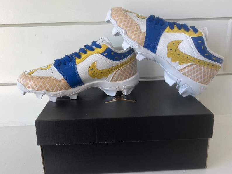 Puede incluir: Un par de tacos blancos, azules y amarillos con un dise&ntilde;o &uacute;nico. Los tacos presentan un patr&oacute;n similar a un gofre en la puntera y el tal&oacute;n, con un logotipo de Nike amarillo. Los zapatos est&aacute;n colocados encima de una caja negra.