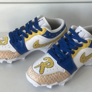 Puede incluir: Par de tacos deportivos blancos y azules con detalles amarillos y cordones azules. Los tacos presentan una "R" dorada en la puntera y un dise&ntilde;o de swoosh dorado. El &aacute;rea de la puntera tiene un patr&oacute;n de gofre.
