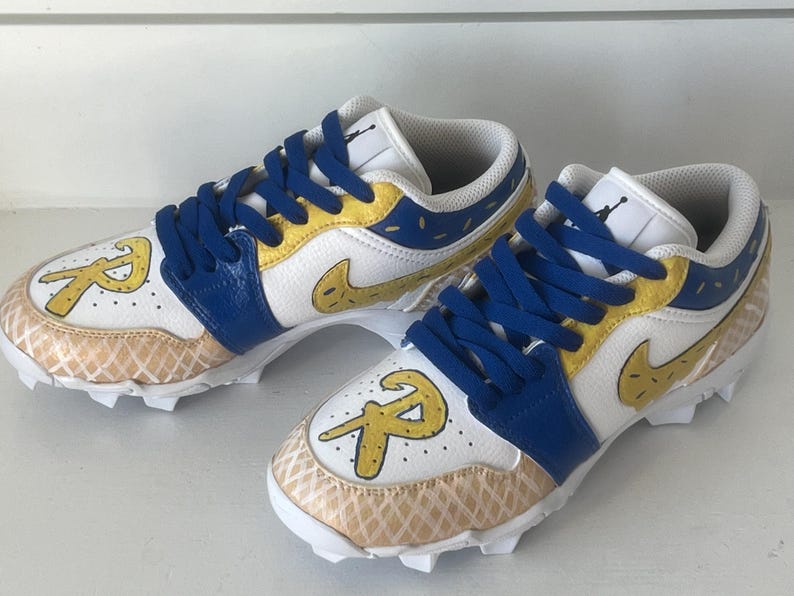 Puede incluir: Par de tacos deportivos blancos y azules con detalles amarillos y cordones azules. Los tacos presentan una "R" dorada en la puntera y un dise&ntilde;o de swoosh dorado. El &aacute;rea de la puntera tiene un patr&oacute;n de gofre.