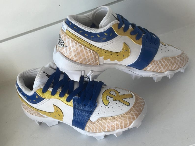 Puede incluir: Par de tacos blancos, azules y amarillos con un dise&ntilde;o &uacute;nico. Los zapatos presentan una correa azul, detalles amarillos y un patr&oacute;n de gofre. El logo de Nike es amarillo. Los zapatos tienen un logo en el lateral y las letras "R" y "R" en la puntera.