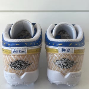Puede incluir: Par de zapatillas Air Jordan personalizadas. Los zapatos presentan una combinaci&oacute;n de colores azul, amarillo y beige con el logotipo de Air Jordan. La palabra "Harkins" est&aacute; escrita en un zapato y "#12" en el otro. Las suelas son blancas.