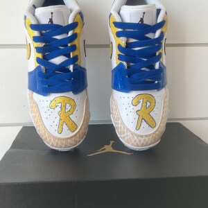 Puede incluir: Par de zapatillas blancas, azules y amarillas con cordones azules. Las zapatillas tienen una "R" amarilla en la puntera y est&aacute;n sobre una caja negra con un logotipo dorado.