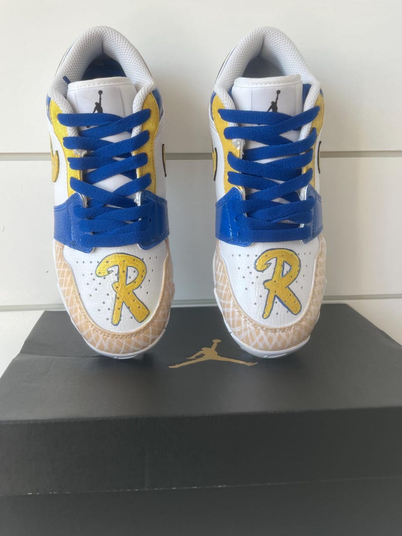 Puede incluir: Par de zapatillas blancas, azules y amarillas con cordones azules. Las zapatillas tienen una "R" amarilla en la puntera y est&aacute;n sobre una caja negra con un logotipo dorado.