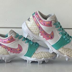 May include: White and teal cleats with a pink and purple sprinkle design. The cleats have a waffle pattern on the sides and a pink swoosh. The laces are teal.