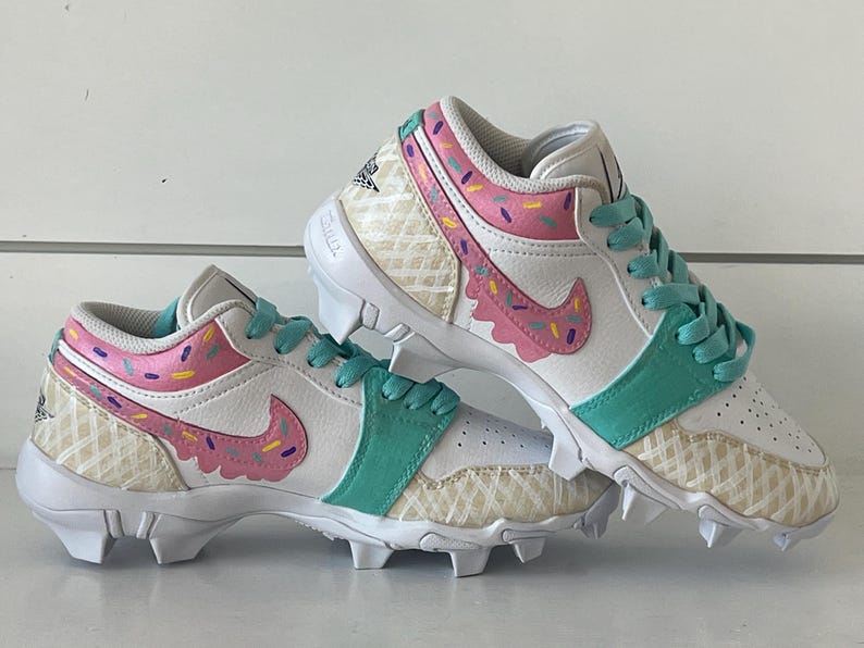 May include: White and teal cleats with a pink and purple sprinkle design. The cleats have a waffle pattern on the sides and a pink swoosh. The laces are teal.