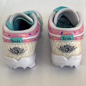 May include: Pair of custom Air Jordan sneakers with a unique ice cream cone design. The shoes feature pink, white, and teal accents, with the names "Brody" and "Irish" on the sides. The soles are white with black cleats.