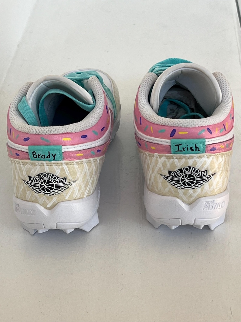 May include: Pair of custom Air Jordan sneakers with a unique ice cream cone design. The shoes feature pink, white, and teal accents, with the names "Brody" and "Irish" on the sides. The soles are white with black cleats.