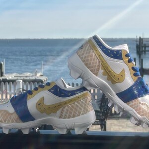 Puede incluir: Un par de tacos blancos, azules y amarillos con un dise&ntilde;o de gofre. Los tacos tienen un logotipo de Nike swoosh amarillo y detalles azules. El fondo muestra una masa de agua y un cielo soleado.