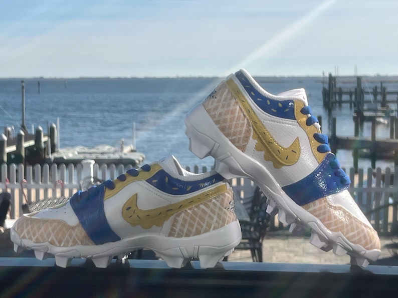 Puede incluir: Un par de tacos blancos, azules y amarillos con un dise&ntilde;o de gofre. Los tacos tienen un logotipo de Nike swoosh amarillo y detalles azules. El fondo muestra una masa de agua y un cielo soleado.