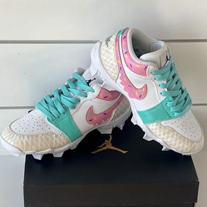 May include: White and teal athletic cleats with a donut design. The cleats feature pink frosting and sprinkles, teal laces, and a gold Jumpman logo. The shoes are on top of a black box with a gold Jumpman logo.