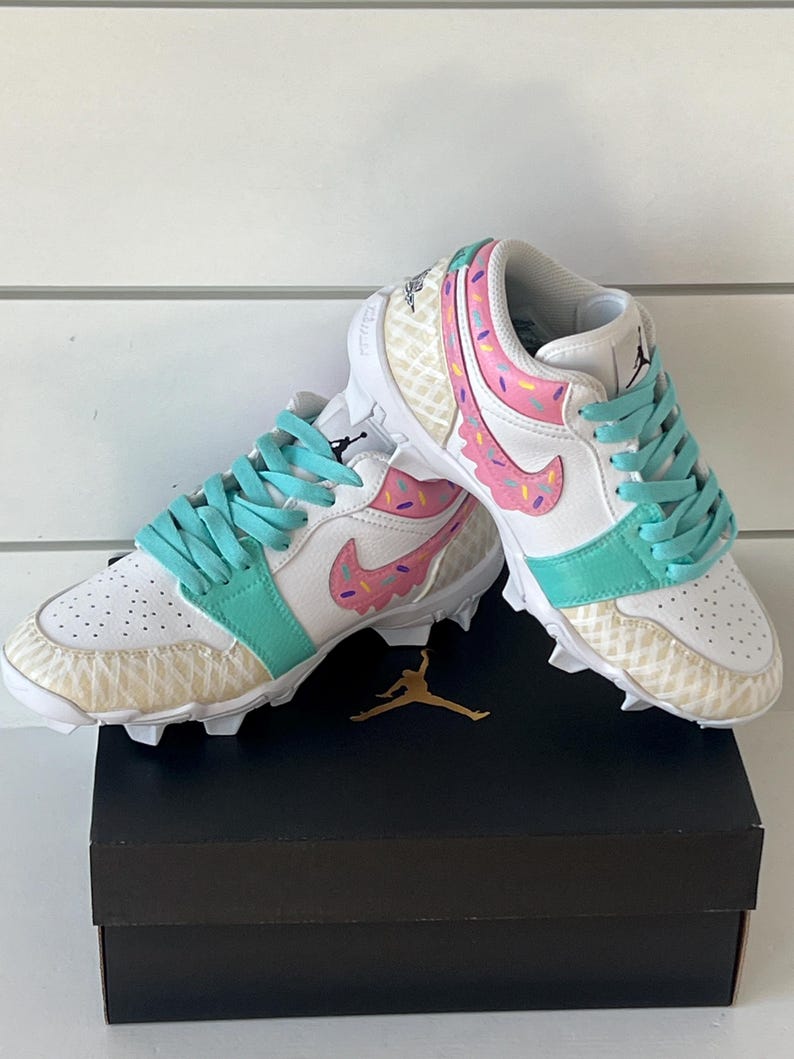 May include: White and teal athletic cleats with a donut design. The cleats feature pink frosting and sprinkles, teal laces, and a gold Jumpman logo. The shoes are on top of a black box with a gold Jumpman logo.