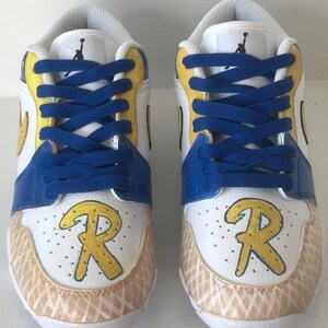 Puede incluir: Par de zapatillas deportivas blancas, azules y amarillas con cordones azules. Las zapatillas presentan la letra "R" amarilla en la puntera y un dise&ntilde;o blanco y beige en la parte delantera inferior. La leng&uuml;eta tiene un logotipo negro.