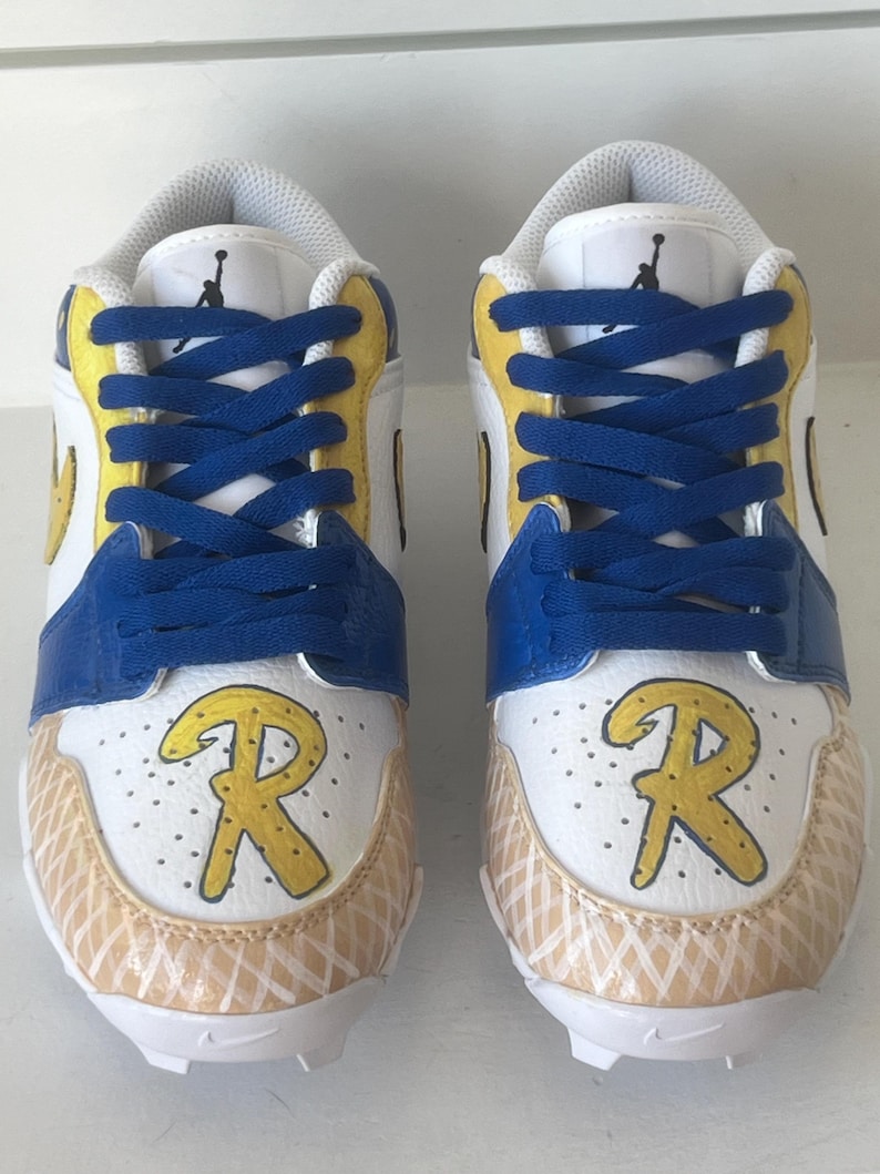 Puede incluir: Par de zapatillas deportivas blancas, azules y amarillas con cordones azules. Las zapatillas presentan la letra "R" amarilla en la puntera y un dise&ntilde;o blanco y beige en la parte delantera inferior. La leng&uuml;eta tiene un logotipo negro.