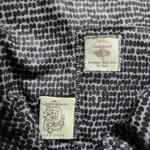 May include: A black and white fabric with a polka dot pattern. Two fabric labels are sewn onto the fabric. The top label is white with red text that reads "Handmade with Love by Kai" and a red mushroom illustration. The bottom label is white with black text that reads "The Whistling Wyvern" and a black and white illustration of a dragon. The text "Handmade" is below the dragon illustration.