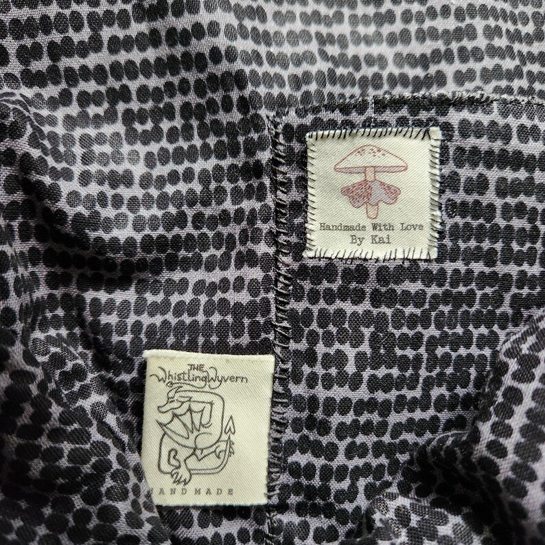 May include: A black and white fabric with a polka dot pattern. Two fabric labels are sewn onto the fabric. The top label is white with red text that reads "Handmade with Love by Kai" and a red mushroom illustration. The bottom label is white with black text that reads "The Whistling Wyvern" and a black and white illustration of a dragon. The text "Handmade" is below the dragon illustration.