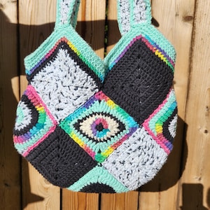 May include: A crocheted tote bag with a patchwork design. The bag is made of black, white, and turquoise yarn with colorful accents. The center of the bag features a large eye design.